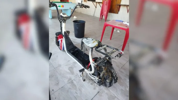 Patinete elétrico danificado, mostrando danos severos na estrutura e no assento, em um ambiente interno com móveis ao fundo.