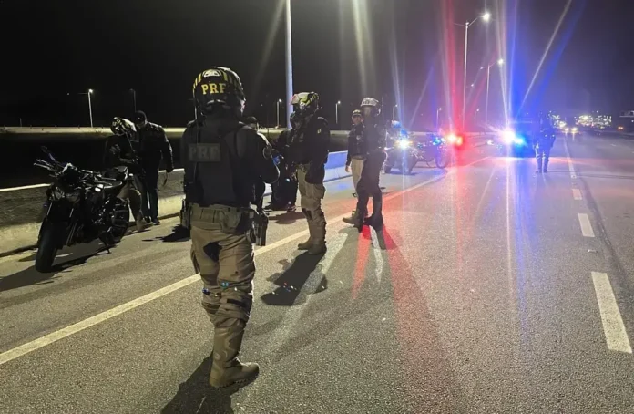 Agentes da Polícia Rodoviária Federal realizando abordagem em motociclistas na rodovia durante a noite.
