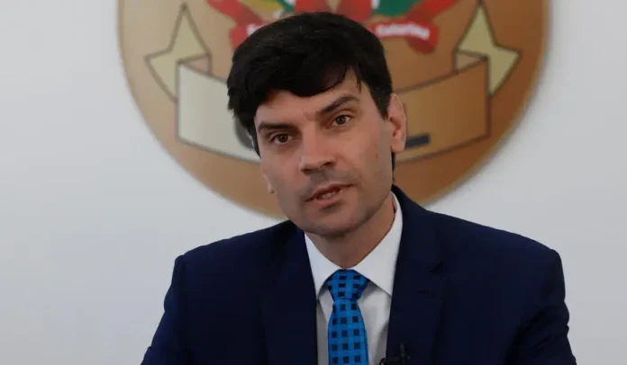 Pessoa em traje formal falando com seriedade, com um emblema ao fundo.