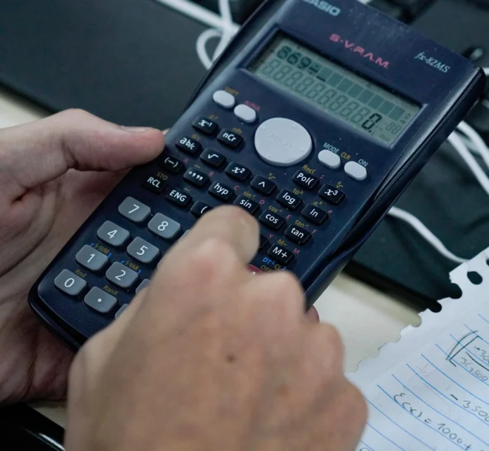 Mão pressionando teclas de uma calculadora científica, com tela exibindo números. Ao fundo, anotações em papel.