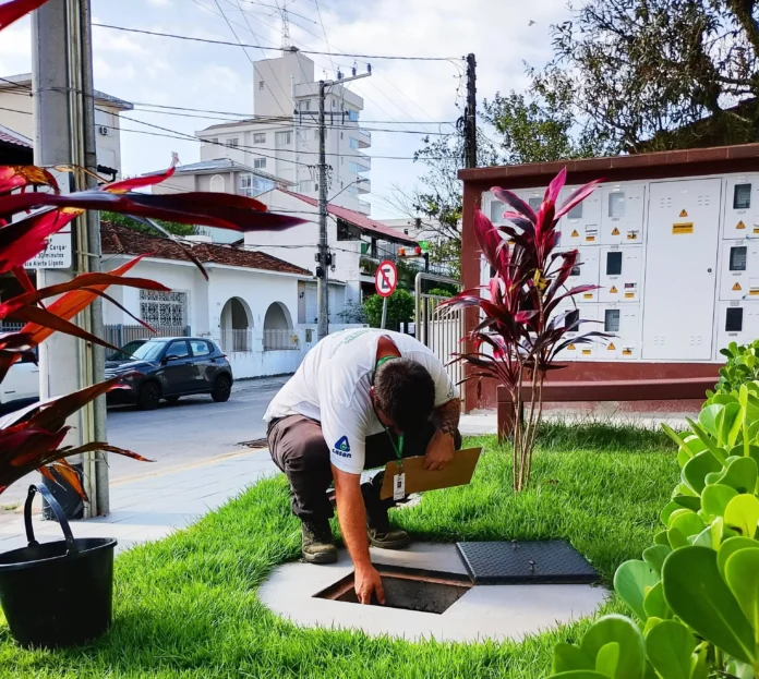 Profissional inspeciona uma tampa de acesso em área ajardinada, com postes e edifícios ao fundo.