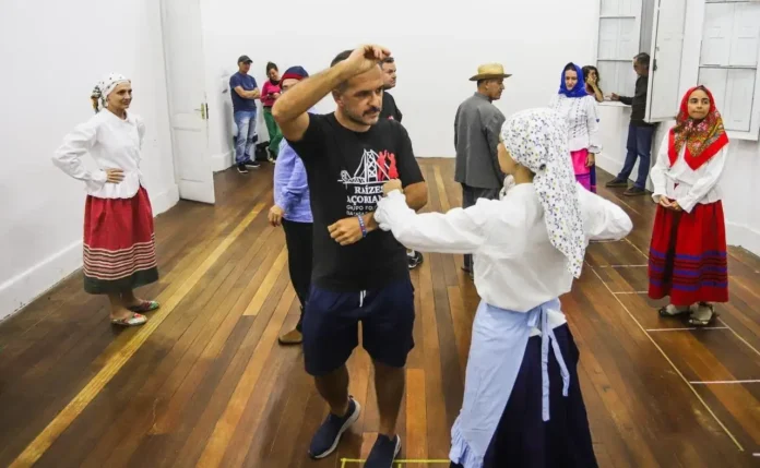 Grupo de pessoas dançando em um ambiente com madeira no chão, vestindo trajes tradicionais.
