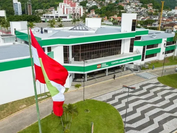 Centro administrativo com fachada verde e janelas grandes, bandeira hasteada em primeiro plano e calçamento em padrão zig-zag.