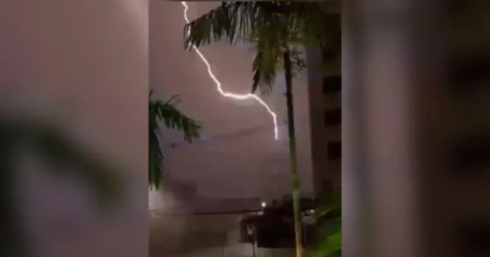 Raio iluminando o céu durante uma tempestade, com árvores e um prédio visíveis ao fundo.