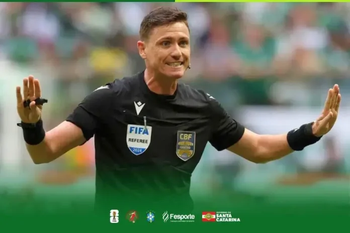 Árbitro em campo, com sinais de decisão, vestindo uniforme preto e insígnias de FIFA e CBF.