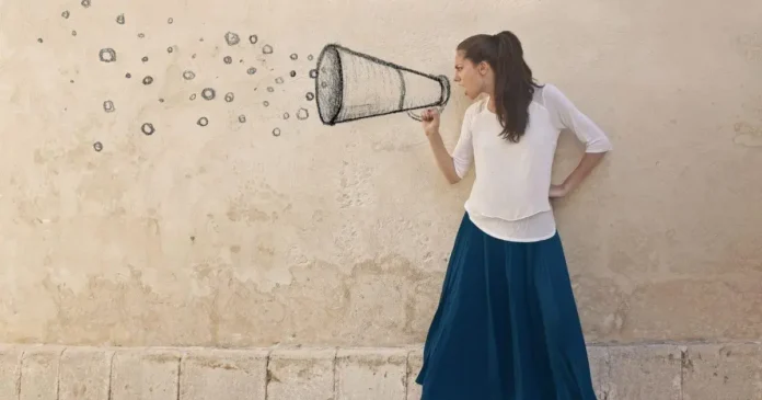 Mulher segurando um megafone estilizado, com um vestido longo azul, em frente a uma parede clara.