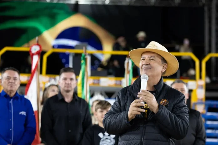 Pessoa em um palco segurando um microfone, com bandeiras ao fundo e uma plateia visível atrás.