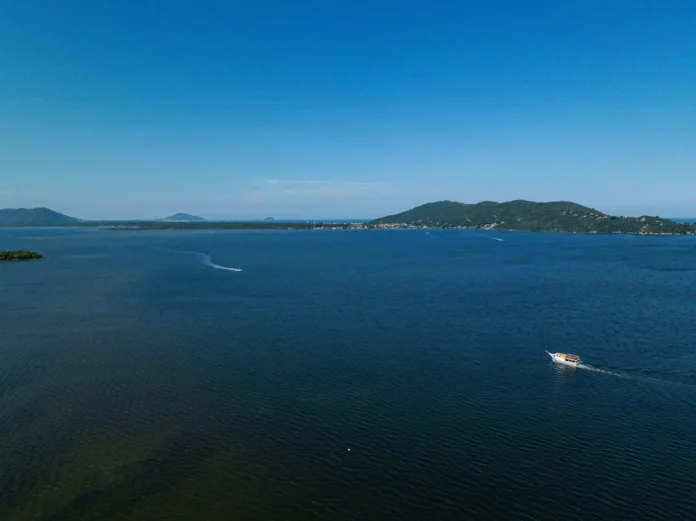 Vista aérea de um vasto corpo d'água com um barco navegando em direção a uma costa montanhosa ao fundo.