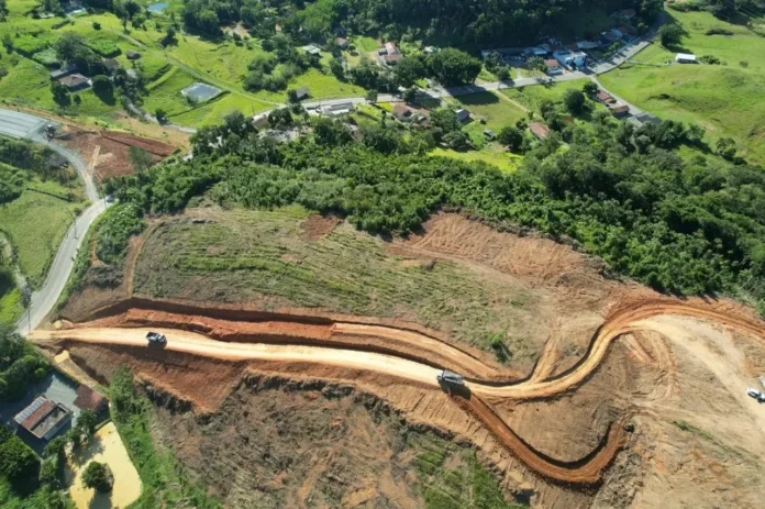 Caminho de terra em construção em área rural, com veículos e vegetação ao redor.