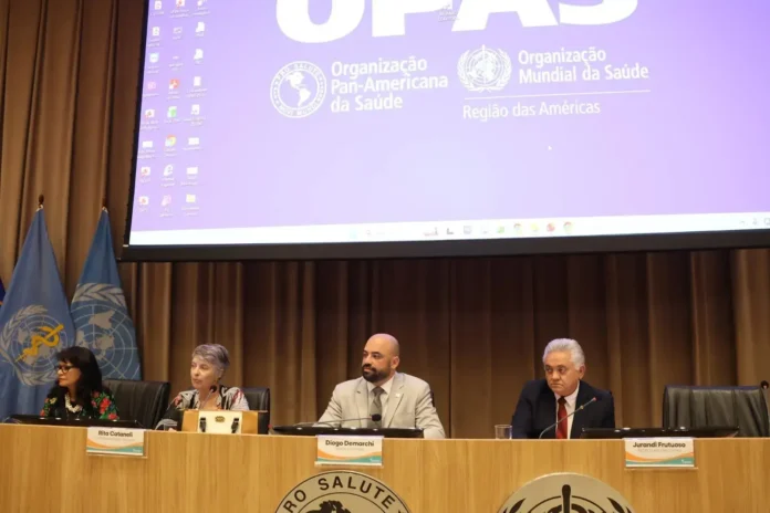 Três pessoas sentadas em uma mesa com logotipo da Organização Pan-Americana da Saúde ao fundo, em um evento oficial.