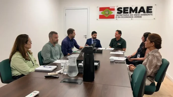 Pessoas em uma reunião sentadas ao redor de uma mesa, com documentos e copos sobre a mesa. O logotipo da Semae está visível na parede.