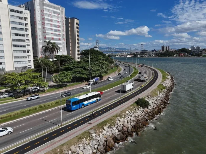 Vista da ciclovia à beira-mar, com veículos na pista e prédios ao fundo, sob céu azul com nuvens.
