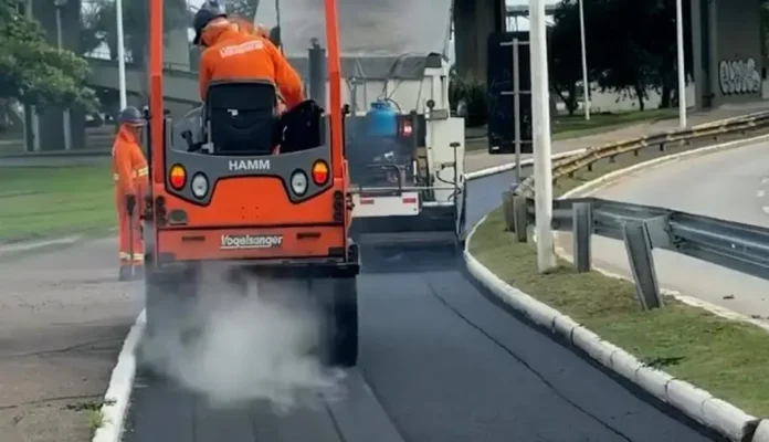 Operadores utilizando maquinário para pavimentação em uma via, com nuvens de fumaça visíveis.
