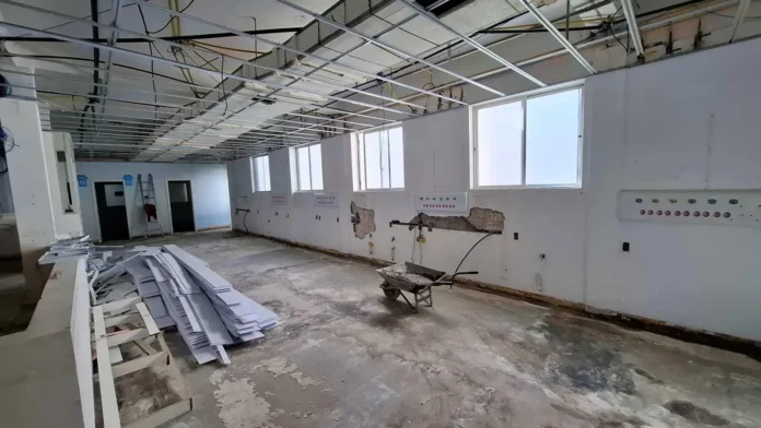 Sala em reforma com pó, estruturas metálicas expostas e carrinho de mão, além de janelas e paredes danificadas.