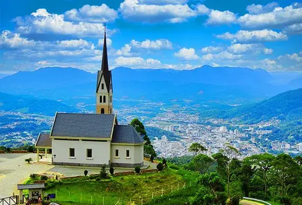 Igreja com torre e telhado escuro em área montanhosa, vista panorâmica de cidades e montanhas ao fundo sob um céu azul com nuvens.
