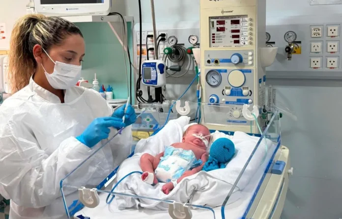 Profissional de saúde cuida de um recém-nascido em uma incubadora, com equipamentos médicos ao fundo.