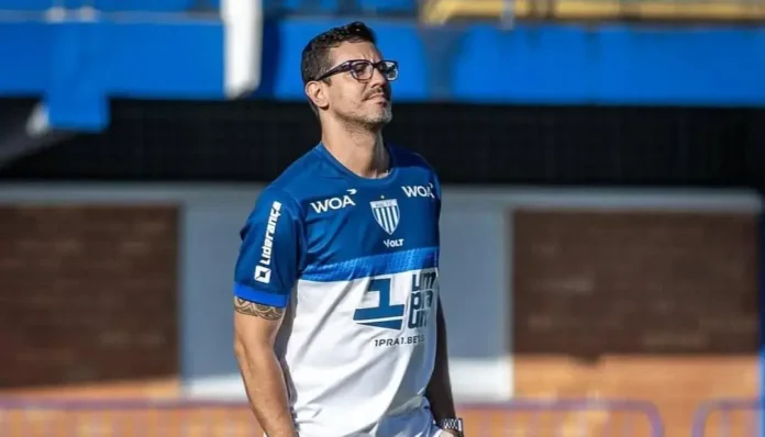 Homem com óculos e camisa esportiva azul e branca, olhando para o campo em ambiente de estádio.