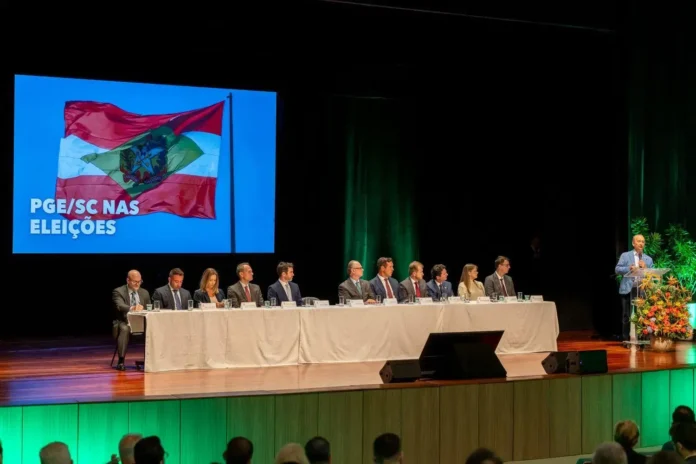 Mesa com diversos indivíduos em um evento, apresentando o seminário "PGE/SC nas Eleições" com um flag em destaque ao fundo.