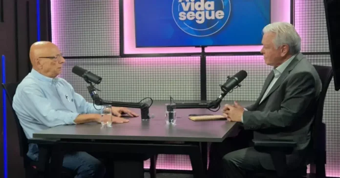 Dois homens em um estúdio conversando, cada um em uma cadeira, com microfones e uma mesa entre eles.