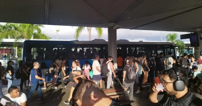Pessoas aguardando ônibus em uma estação de transporte público, com veículo estacionado e sinalização visível.