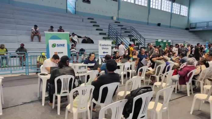 Espaço do evento com mesas de atendimento, participantes sentados em cadeiras brancas e banners do Sine e Cadastro Único.