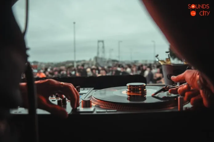 Mãos controlando um toca-discos em um evento, com uma multidão visível ao fundo.
