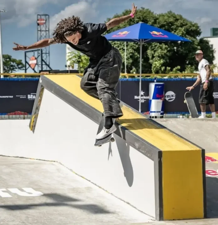 Skatista realiza manobra em uma estrutura inclinada, com céu azul e público ao fundo.