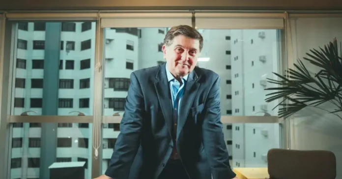 Homem com blazer escuro sorrindo em ambiente interno, próximo a janelas com vista para prédios.