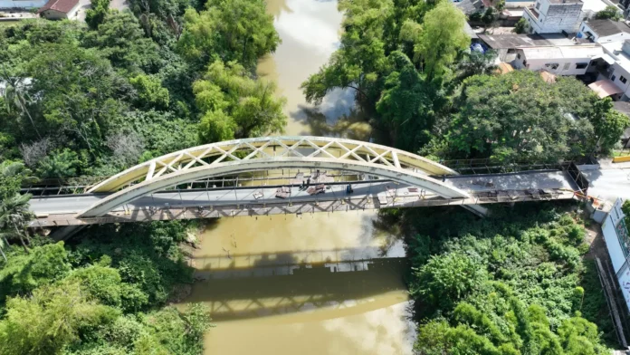 Ponte sobre um rio, rodeada de vegetação, com obras em andamento na estrutura e no pavimento.