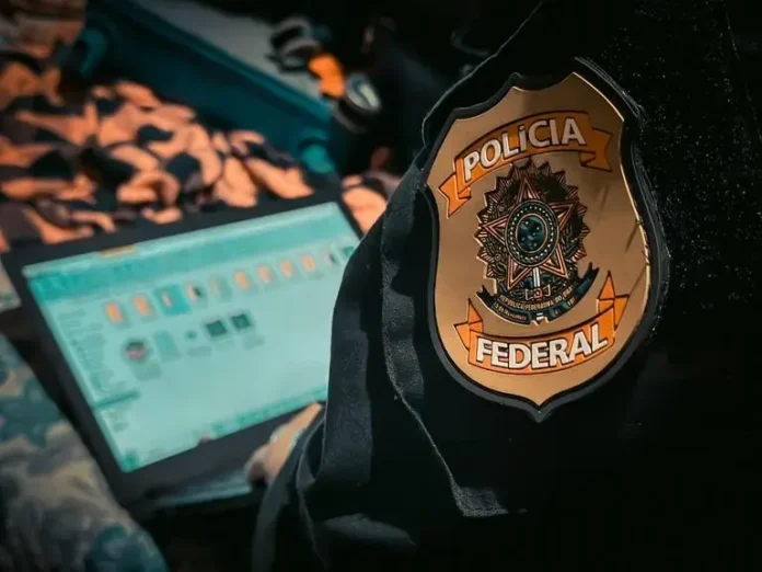 Insígnia da Polícia Federal em uniforme, ao lado de um laptop com tela visível.