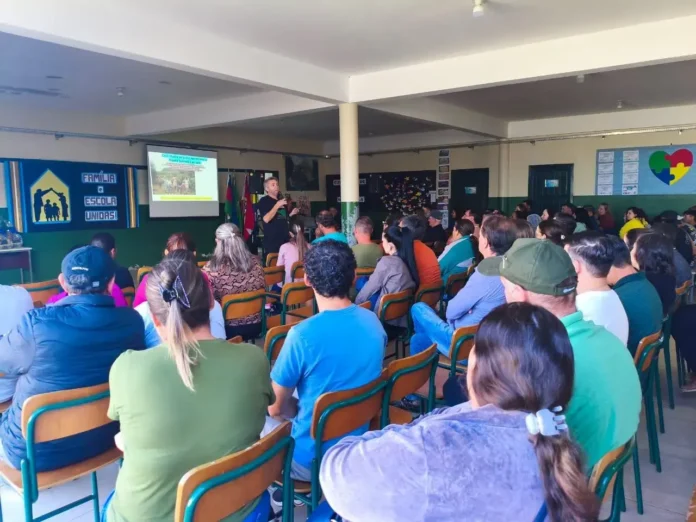 Sala de aula com várias pessoas assistindo a uma apresentação, em frente a um telão com slides e bandeiras.
