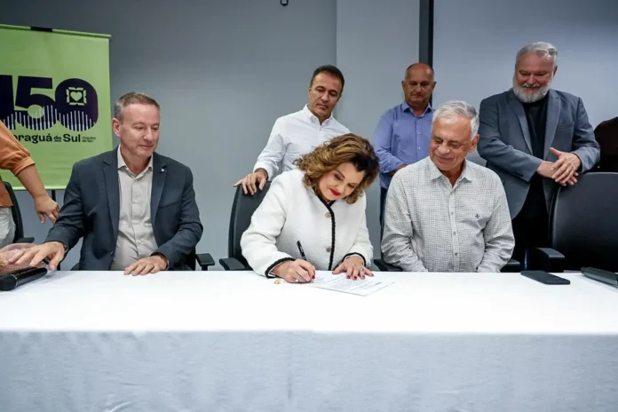 Vice-governadora assina documento em evento, com pessoas ao fundo e uma bandeira ao lado.